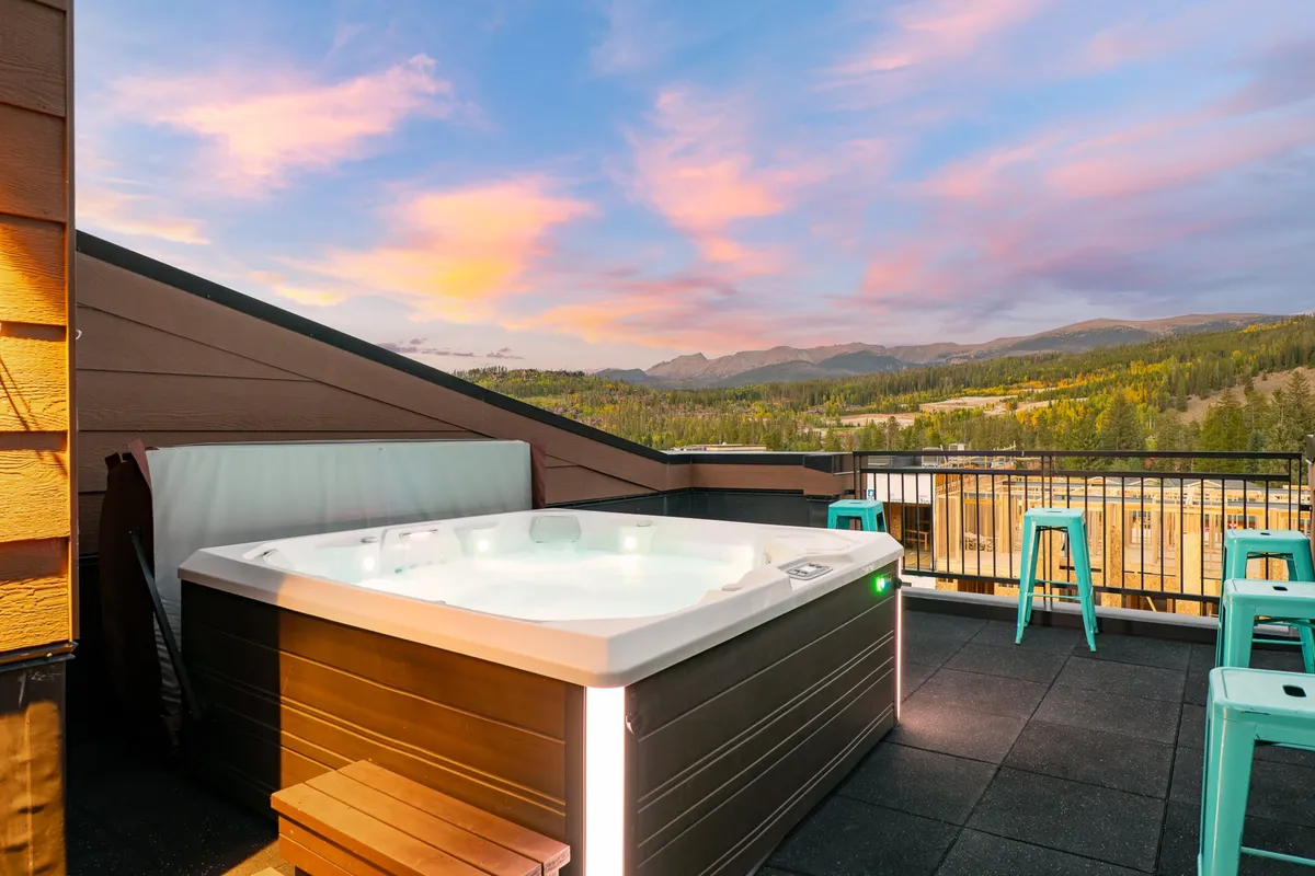 This exterior scene features a custom hot tub on a rooftop patio with sweeping mountain views and dramatic sky colors at sunset. The hot tub is surrounded by inviting seating, creating a picturesque setting for relaxation and social gatherings.