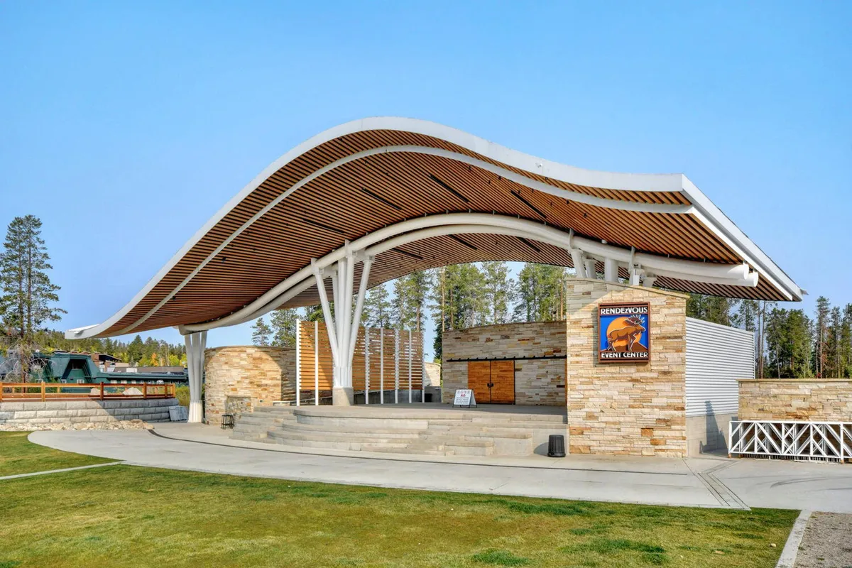 An outdoor amphitheater with a distinctive curved roof, stone facade, and steps leading up to a stage, set against a backdrop suitable for concerts or community events. The space is vibrant and inviting, ideal for gatherings and celebrations.