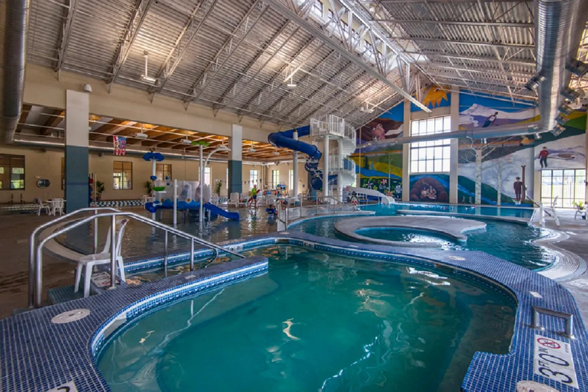 A bright, playful indoor aquatic center featuring a large swimming pool, water slide, and interactive water play features. Children and families are visible enjoying the space, with colorful murals adding to the cheerful atmosphere.