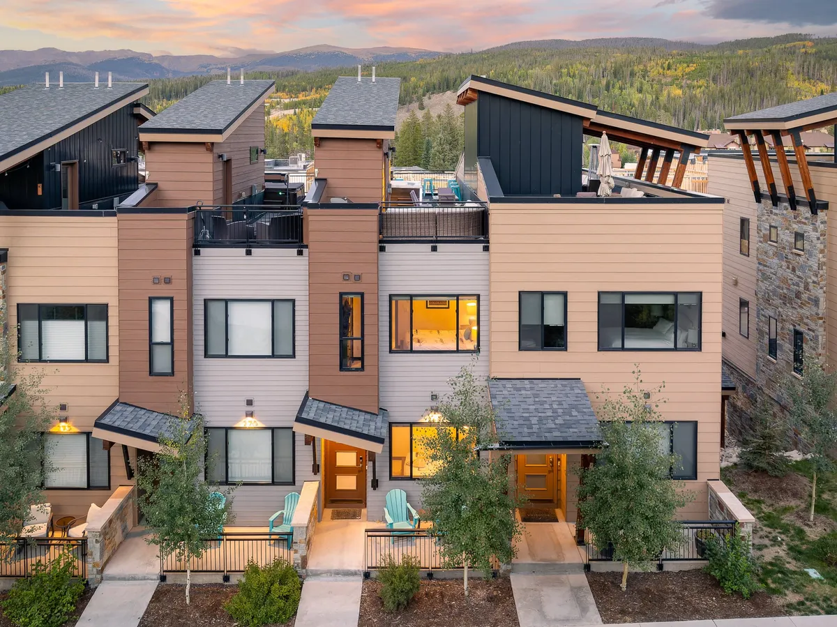 The image showcases a modern single-family villa with striking architecture, a beautifully landscaped yard, and a spacious outdoor seating area perfect for gatherings. The balcony glows with sunset hues while guests relax by a fire pit and enjoy sweeping mountain views, evoking a sense of warmth and togetherness.