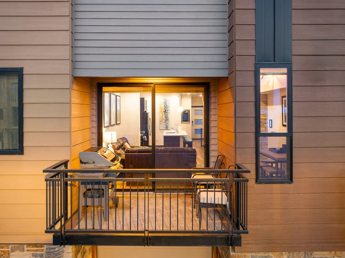 A spacious balcony overlooking a warmly lit living room, with stylish furniture and ambient lighting visible through large windows. The balcony offers a seamless transition between indoor comfort and outdoor enjoyment, ideal for relaxing or entertaining.