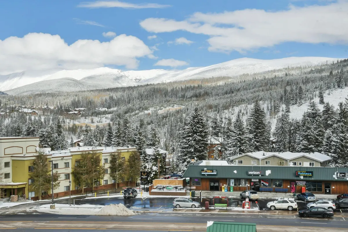A panoramic perspective of snow-covered mountains rising above pine trees and vibrant commercial buildings, suggesting a blend of natural beauty and bustling community life. This scene hints at après-ski activities, shopping, and dining in the local area.