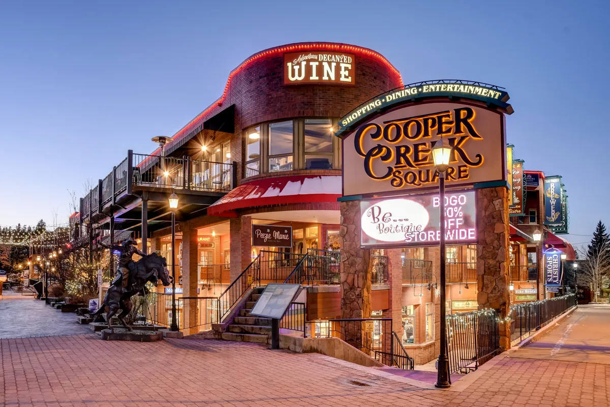 The image features the Cooper Creek Square sign with a wine shop entrance and illuminated outdoor seating, capturing the energy of downtown Winter Park in the evening. The area is alive with people enjoying dining, shopping, and live entertainment.