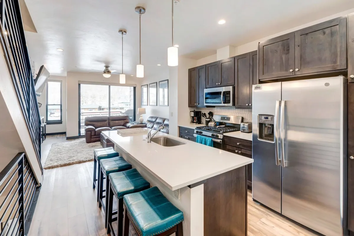 This image features a beautiful, contemporary kitchen with a large island as its centerpiece. Stainless steel appliances glisten beneath ample natural light streaming in from large windows. Comfortable seating at the island and in an adjacent lounge area suggests a space ideal for both cooking and socializing. The overall design is welcoming, stylish, and functional—perfect for family gatherings or casual breakfasts.