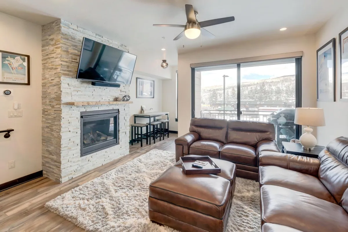 This inviting living room features a leather sectional sofa, a central fireplace, large windows with mountain views, and an HD television mounted above the fire. The space is flooded with natural light, and the coffee table invites board games or relaxed conversation.