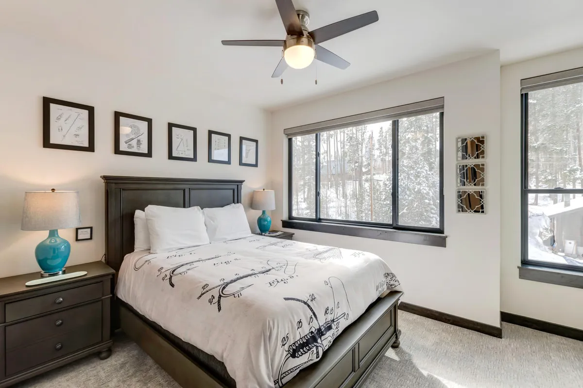 A tranquil bedroom with a plush bed covered in patterned bedding, large windows that allow natural light to flood in, decorative wall art, and a nightstand with a lamp. The space feels cozy, private, and thoughtfully designed for rest.