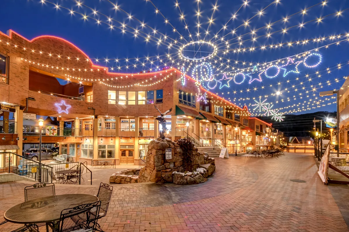 String lights hang overhead, decorative stars glimmer, and a stone fountain forms the centerpiece of a lively outdoor plaza. People are gathered for community events, enjoying outdoor dining and shopping under the festive lights.
