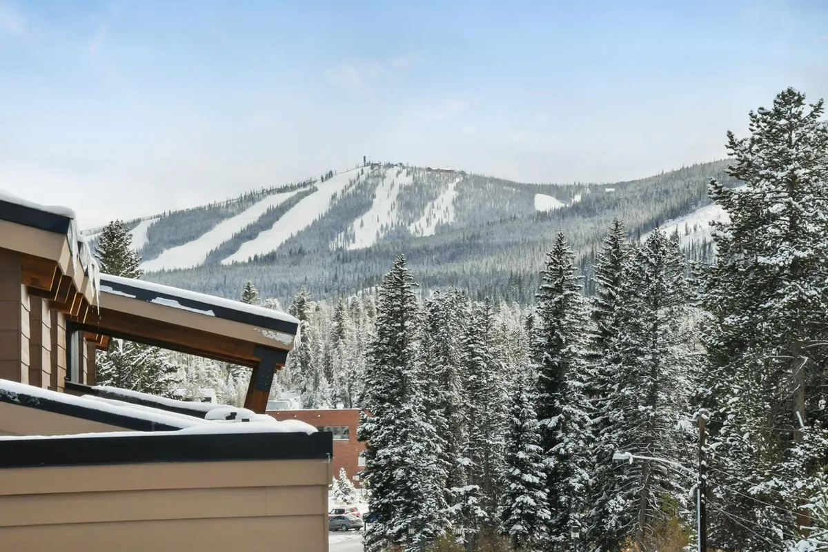 A breathtaking view of snow-covered mountains framed by tall pine trees, with visible ski slopes winding down the mountainside. The scene exudes a sense of tranquility and adventure, highlighting the region’s winter charm and access to outdoor recreation.